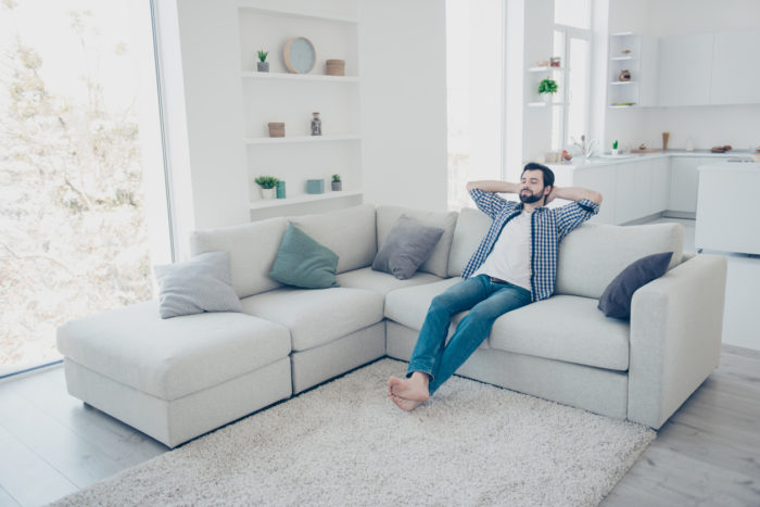 Full-size portrait of meditative trendy single man with stubble in jeans shirt sitting on sofa in modern livingroom holding hands behind the head with close eyes enjoying free time